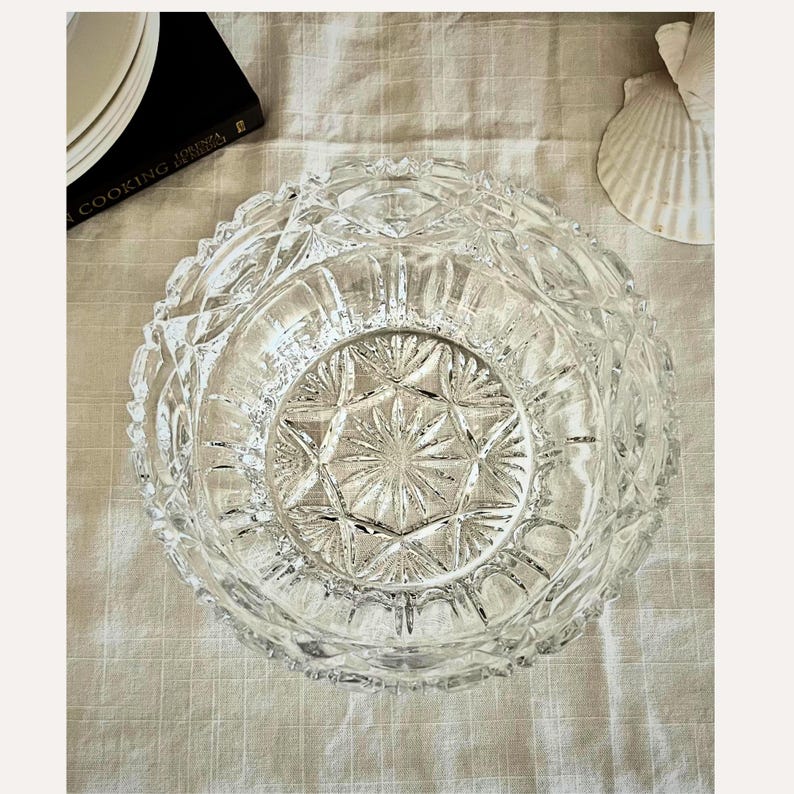 Peut inclure: Un bol en verre taill&eacute; transparent avec un motif en &eacute;toile au centre et un motif taill&eacute; en diamant sur le bord. Le bol repose sur une surface textur&eacute;e de couleur claire. Un livre de cuisine et un coquillage sont partiellement visibles en arri&egrave;re-plan.