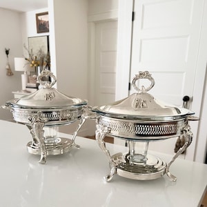 May include: Two silver-plated chafing dishes with clear glass inserts, ornate pierced metal sides, and decorative lids featuring floral finials. Each dish is supported by a pedestal base with four curved legs and includes a central candle holder.