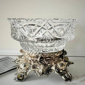 Peut inclure: Un saladier en verre taill&eacute; transparent avec un motif de losanges repose sur un socle en m&eacute;tal argent&eacute; orn&eacute;. La base pr&eacute;sente des volutes complexes et trois pieds d&eacute;coratifs. Un tissu ray&eacute; est visible.