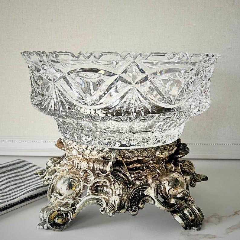 Peut inclure: Un saladier en verre taill&eacute; transparent avec un motif de losanges repose sur un socle en m&eacute;tal argent&eacute; orn&eacute;. La base pr&eacute;sente des volutes complexes et trois pieds d&eacute;coratifs. Un tissu ray&eacute; est visible.