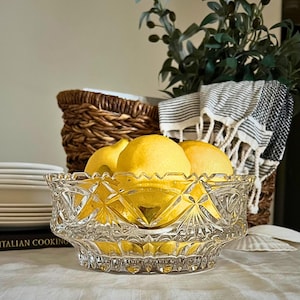 Peut inclure: Un bol en verre taill&eacute; transparent rempli de citrons jaunes. Le bol pr&eacute;sente un motif g&eacute;om&eacute;trique. En arri&egrave;re-plan, un panier en osier, une serviette ray&eacute;e et de la verdure composent une nature morte. Des assiettes blanches sont visibles.