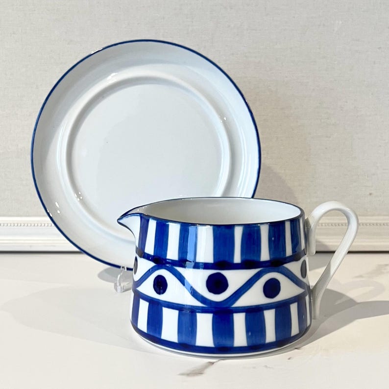 Op de afbeelding: Witte keramische kan met een blauw-wit gestreept en gestippeld patroon, en een bijpassend wit bord met een blauwe rand. De kan heeft een handvat en een schenktuit.