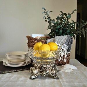 Peut inclure: Un compotier en verre taill&eacute; transparent rempli de citrons jaunes, pos&eacute; sur un socle argent&eacute; orn&eacute;. Une pile d'assiettes et de bols blancs, un panier en osier et un livre intitul&eacute; "THE RENAISSANCE OF ITALIAN COOKING" sont &eacute;galement visibles.