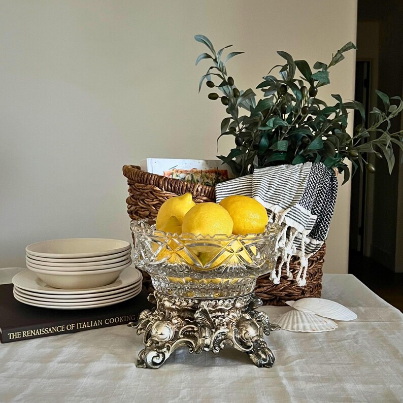 Peut inclure: Un compotier en verre taill&eacute; transparent rempli de citrons jaunes, pos&eacute; sur un socle argent&eacute; orn&eacute;. Une pile d'assiettes et de bols blancs, un panier en osier et un livre intitul&eacute; "THE RENAISSANCE OF ITALIAN COOKING" sont &eacute;galement visibles.