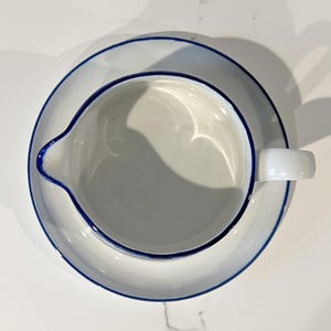 Op de afbeelding: Witte keramische melkkan en schotel set met een blauwe rand. De melkkan heeft een tuit en een handvat en staat op een bijpassende schotel. De set is schoon en eenvoudig, perfect voor het serveren van room of melk.