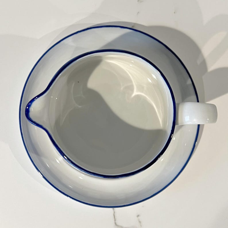 Op de afbeelding: Witte keramische melkkan en schotel set met een blauwe rand. De melkkan heeft een tuit en een handvat en staat op een bijpassende schotel. De set is schoon en eenvoudig, perfect voor het serveren van room of melk.