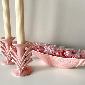 May include: Two pink speckled candle holders with cream-colored candles and a pink speckled boat-shaped dish filled with wrapped red and white peppermint candies. The candle holders have a decorative leaf design.
