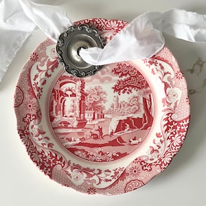 May include: A red and white decorative plate with a landscape scene, a silver napkin ring, and a white ribbon. The plate has a detailed floral border and a central image of a classical landscape. The napkin ring is silver and holds a white ribbon.