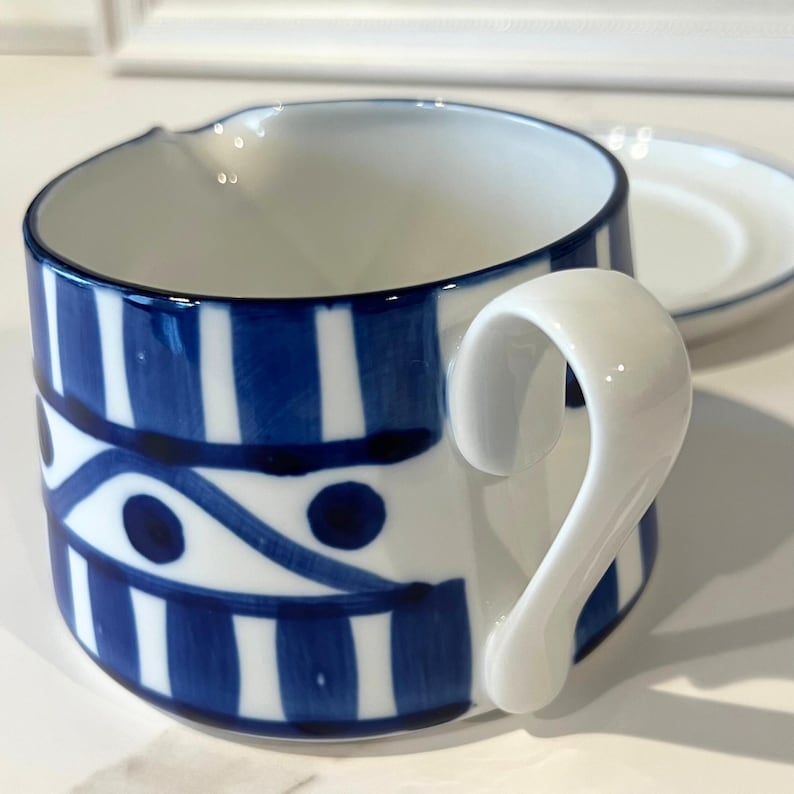 Op de afbeelding: Een witte keramische beker met een blauw-wit gestreept patroon en een bijpassende schotel. De beker heeft een uniek handvat en een schenktuit. Het ontwerp bevat blauwe strepen en cirkelvormige accenten.