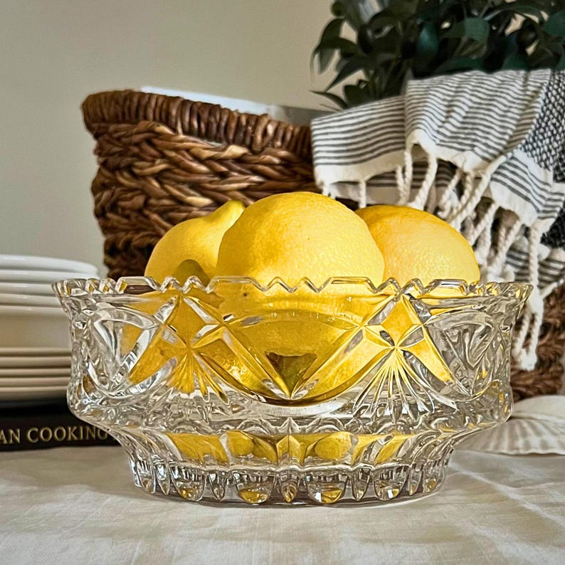 Peut inclure: Un bol en verre taill&eacute; transparent rempli de citrons jaunes. Le bol pr&eacute;sente un motif en losanges et est pos&eacute; sur une surface blanche. Un panier en osier et un torchon &agrave; rayures sont visibles en arri&egrave;re-plan, sugg&eacute;rant une ambiance de cuisine.