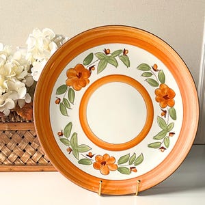 May include: A decorative ceramic plate with an orange rim and a central floral design. The plate features orange flowers and green leaves arranged around the center. The plate is displayed on a gold stand.