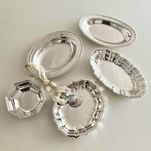 May include: A collection of four polished silver-toned metal trays in various shapes, including oval and octagonal designs, accompanied by a delicate white beaded necklace featuring a small, iridescent seashell pendant resting on one tray.