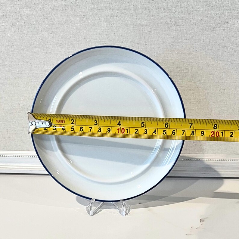 Op de afbeelding: Witte bord met een blauwe rand, ongeveer 20 cm in diameter. Het bord wordt gepresenteerd op een transparante standaard, met een meetlint. Het bord heeft een eenvoudig, klassiek ontwerp.