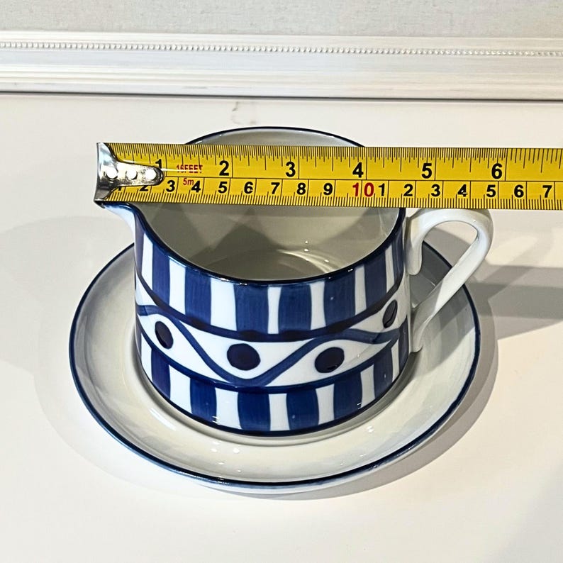 Op de afbeelding: Een witte en blauwe keramische juskom met een bijpassende schotel. De juskom heeft een blauw-wit gestreept patroon met een golvend ontwerp. De juskom meet ongeveer 7,6 cm hoog en 15,2 cm lang.