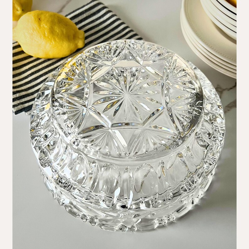 Peut inclure: Un bol en verre taill&eacute; transparent avec un couvercle, pr&eacute;sentant un motif en forme d'&eacute;clat d'&eacute;toile. Le bol est rond avec un bord d&eacute;coratif. Deux citrons jaunes et un tissu &agrave; rayures noires et blanches sont en arri&egrave;re-plan.