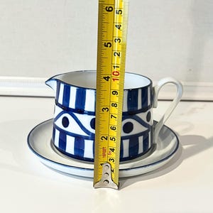Op de afbeelding: Een witte en blauwe keramische juskom met bijpassende schotel. De juskom heeft een handvat en een tuit. Het ontwerp heeft blauwe strepen en oogachtige patronen. De hoogte is ongeveer 10 cm.