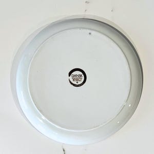 Op de afbeelding: Witte keramische plaat met een grijze rand. De onderkant heeft het "DANSK INTERNATIONAL DESIGNS PHILIPPINES" logo in een zwarte cirkel. De plaat heeft een eenvoudig, modern ontwerp.