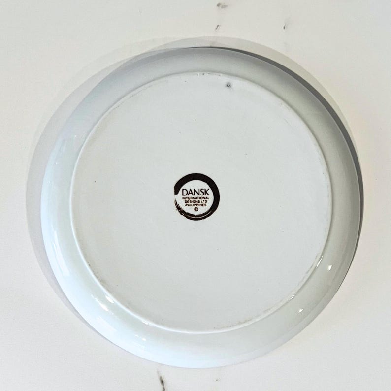 Op de afbeelding: Witte keramische plaat met een grijze rand. De onderkant heeft het "DANSK INTERNATIONAL DESIGNS PHILIPPINES" logo in een zwarte cirkel. De plaat heeft een eenvoudig, modern ontwerp.