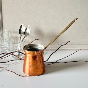 May include: A copper cezve coffee pot with a long brass handle. The pot has a rounded base and a narrow top. Two silver spoons and small glasses are in the background. Decorative twigs are in the foreground.