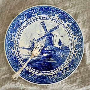May include: A blue and white ceramic plate with a windmill scene and floral border. A silver fork rests on the plate. The plate's design features windmills, water, and a detailed border. The plate is a decorative item.