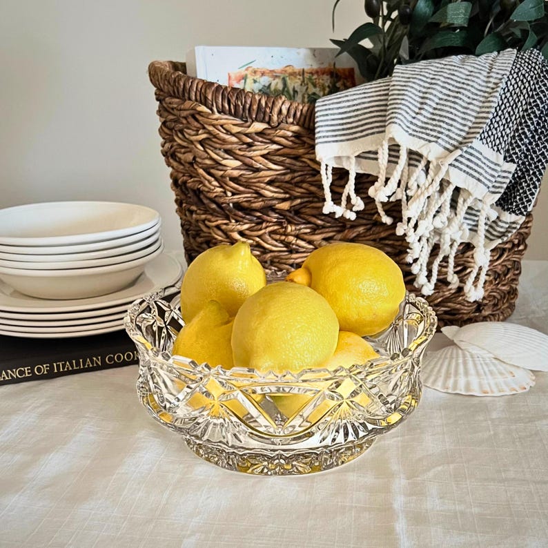 Peut inclure: Un bol en cristal clair rempli de citrons jaune vif. Le bol est pos&eacute; sur une nappe blanche, avec une pile d'assiettes blanches et un panier en osier en arri&egrave;re-plan. Une serviette &agrave; rayures noires et blanches est drap&eacute;e sur le panier.