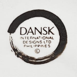Op de afbeelding: Een close-up van een wit oppervlak met een zwart cirkelvormig ontwerp. Het ontwerp bevat het woord "DANSK" in een vetgedrukte letter, met daaronder "INTERNATIONAL DESIGNS LTD PHILIPPINES". Er is ook een copyrightsymbool aanwezig.