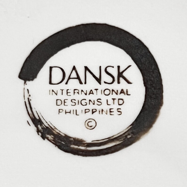 Op de afbeelding: Een close-up van een wit oppervlak met een zwart cirkelvormig ontwerp. Het ontwerp bevat het woord "DANSK" in een vetgedrukte letter, met daaronder "INTERNATIONAL DESIGNS LTD PHILIPPINES". Er is ook een copyrightsymbool aanwezig.