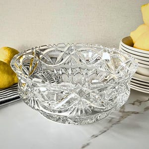 Peut inclure: Un saladier en verre taill&eacute; transparent avec un motif en &eacute;toile et g&eacute;om&eacute;trique. Le bol a un bord festonn&eacute; et repose sur un petit socle. Des citrons et une pile d'assiettes blanches sont en arri&egrave;re-plan.