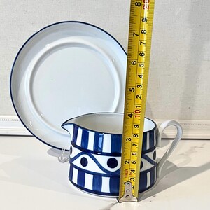 Op de afbeelding: Een witte keramische creamer met een blauw-wit geometrisch patroon, een handvat en een tuit, naast een witte plaat met een blauwe rand. De creamer is ongeveer 7.6 cm hoog.