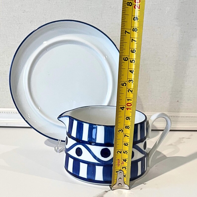 Op de afbeelding: Een witte keramische creamer met een blauw-wit geometrisch patroon, een handvat en een tuit, naast een witte plaat met een blauwe rand. De creamer is ongeveer 7.6 cm hoog.