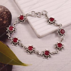 Puede incluir: Un brazalete de plata con una serie de piedras preciosas redondas de color rojo intenso engastadas en marcos en forma de estrella. El brazalete se exhibe sobre una superficie blanca, mostrando su diseño.