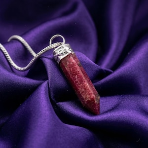 Może przedstawiać: Srebrny naszyjnik z wisiorkiem z głęboko czerwonym, fasetowanym kamieniem szlachetnym. Wisiorek ma podłużny, sześciokątny kształt ze srebrną nakładką. Naszyjnik spoczywa na fioletowej satynie.