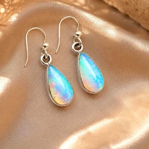 May include: A pair of silver drop earrings featuring teardrop-shaped iridescent opals. Each earring has a curved silver hook wire attached to a small circular ring, which connects to the smooth, polished, pear-shaped opal gemstone setting.