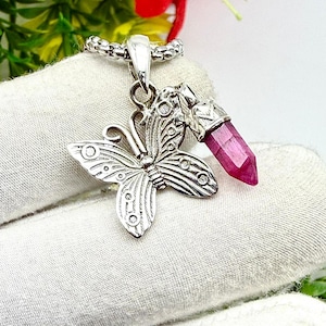 May include: A silver butterfly pendant with a faceted, pink gemstone. The butterfly has detailed wing patterns and is attached to a chain. The gemstone is a deep pink color and is cut into a pointed shape.