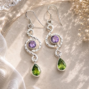 May include: A pair of silver dangle earrings featuring a hammered, swirling metal design. Each earring is set with a round, faceted purple amethyst stone in the centre and a pear-shaped, faceted green peridot stone hanging at the bottom.
