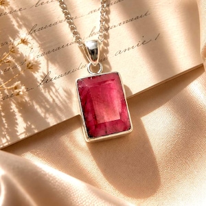 May include: A rectangular, deep pink gemstone pendant set in a polished silver frame, suspended from a matching silver chain. The stone features a faceted surface with visible natural inclusions, reflecting light across its vibrant, saturated red-pink hue.