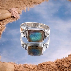 May include: A sterling silver ring featuring a square-cut labradorite gemstone with iridescent blue and green flashes. The band has a polished metallic finish and includes the hallmark 925 stamped on the interior surface of the silver metal band.