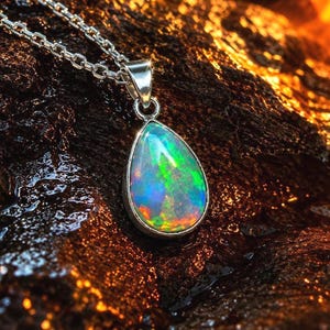 Könnte beinhalten: Ein tropfenförmiger Opal-Anhänger in einer polierten Silberfassung, der an einer passenden Silberkette hängt. Der Opal zeigt leuchtende grüne, blaue und orangefarbene Feuerreflexe auf seiner gesamten Oberfläche und reflektiert das Licht.