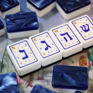 May include: A set of rectangular mahjong tiles with white faces and dark blue marbled backs. Four tiles are arranged in a row, each featuring a blue Hebrew letter, a small gold letter N, E, W, or S in the corner, and a pattern of small gold stars.