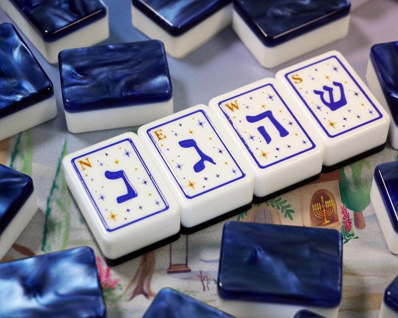 May include: A set of rectangular mahjong tiles with white faces and dark blue marbled backs. Four tiles are arranged in a row, each featuring a blue Hebrew letter, a small gold letter N, E, W, or S in the corner, and a pattern of small gold stars.