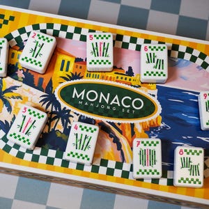 Op de afbeelding: Een rechthoekige Monaco Mahjong-speldoos met een kleurrijke illustratie van een kustlandschap met palmbomen en gebouwen. Negen witte rechthoekige tegels met groene bamboepatronen en een roze flamingo zijn op het deksel gerangschikt.