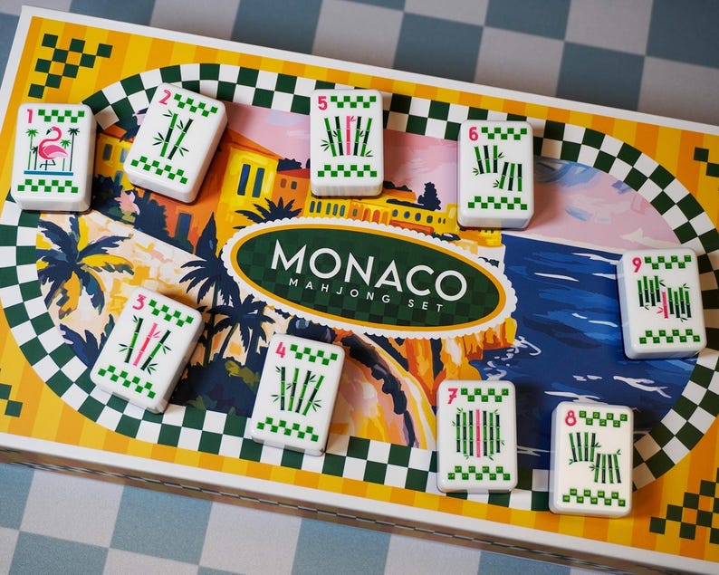 Op de afbeelding: Een rechthoekige Monaco Mahjong-speldoos met een kleurrijke illustratie van een kustlandschap met palmbomen en gebouwen. Negen witte rechthoekige tegels met groene bamboepatronen en een roze flamingo zijn op het deksel gerangschikt.