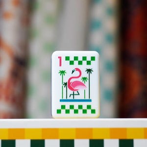 Op de afbeelding: Een rechthoekige witte speltiegel met een illustratie van een roze flamingo in het midden tussen groene palmbomen. De tegel toont het cijfer &eacute;&eacute;n in het roze in de linkerbovenhoek, met groene geblokte randen aan de boven- en onderkant.