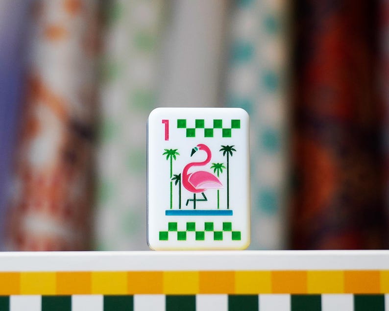 Op de afbeelding: Een rechthoekige witte speltiegel met een illustratie van een roze flamingo in het midden tussen groene palmbomen. De tegel toont het cijfer &eacute;&eacute;n in het roze in de linkerbovenhoek, met groene geblokte randen aan de boven- en onderkant.