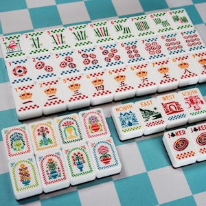 Op de afbeelding: Een set witte rechthoekige mahjongtegels met kleurrijke illustraties van bloemen, kronen, bamboe, cijfers en windstreken. De tegels tonen diverse symbolen waaronder een flamingo, roulettewielen en labels zoals North, East, West, South en Joker.