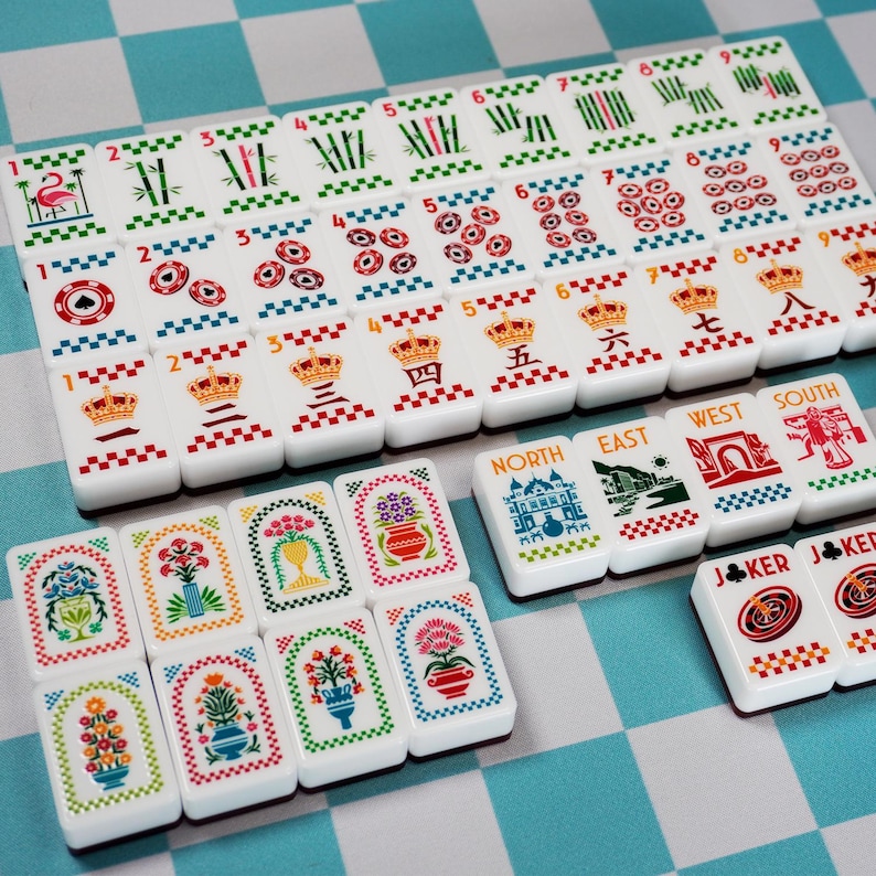 Op de afbeelding: Een set witte rechthoekige mahjongtegels met kleurrijke illustraties van bloemen, kronen, bamboe, cijfers en windstreken. De tegels tonen diverse symbolen waaronder een flamingo, roulettewielen en labels zoals North, East, West, South en Joker.