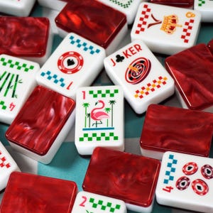 Op de afbeelding: Een verzameling rechthoekige mahjongstenen met witte voorzijden en kleurrijke prints, waaronder een roze flamingo, een joker, een kroon en diverse symbolen, gecombineerd met donkerrode, gemarmerde, doorschijnende achterzijden.