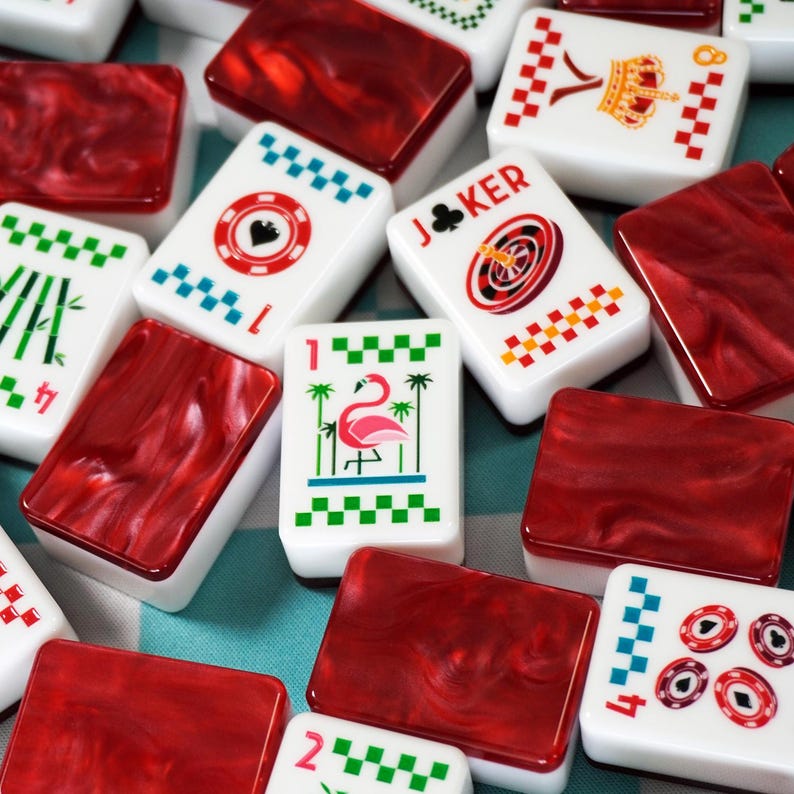 Op de afbeelding: Een verzameling rechthoekige mahjongstenen met witte voorzijden en kleurrijke prints, waaronder een roze flamingo, een joker, een kroon en diverse symbolen, gecombineerd met donkerrode, gemarmerde, doorschijnende achterzijden.