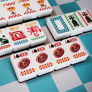 Op de afbeelding: Een verzameling witte rechthoekige mahjongtegels met kleurrijke afbeeldingen, waaronder rode kronen, raceauto's, windstreken zoals West en South, en vier Joker-tegels met roulettewielen en de tekst JOKER met klaver-symbolen.