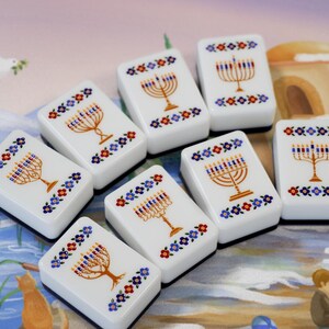 May include: A set of seven white rectangular tiles, each featuring a gold menorah illustration with blue and red candles, surrounded by a decorative border of small blue, red, and yellow flowers. The tiles have a smooth, glossy finish.